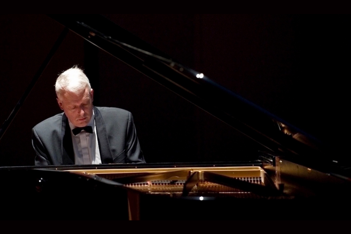 Cette image montre un homme en costume noir en train de jouer du piano à queue sur une scène. Il semble très concentré, la tête légèrement baissée vers les touches du piano. L’éclairage sombre met en valeur le musicien et l’instrument, créant une ambiance élégante et artistique.
