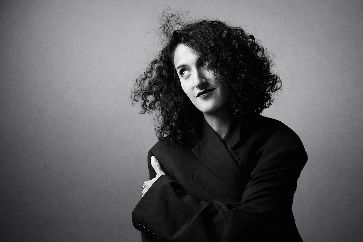 Portrait en noir et blanc d’une femme aux cheveux bouclés, vêtue d’une veste sombre, regardant légèrement vers le haut sur un fond neutre.