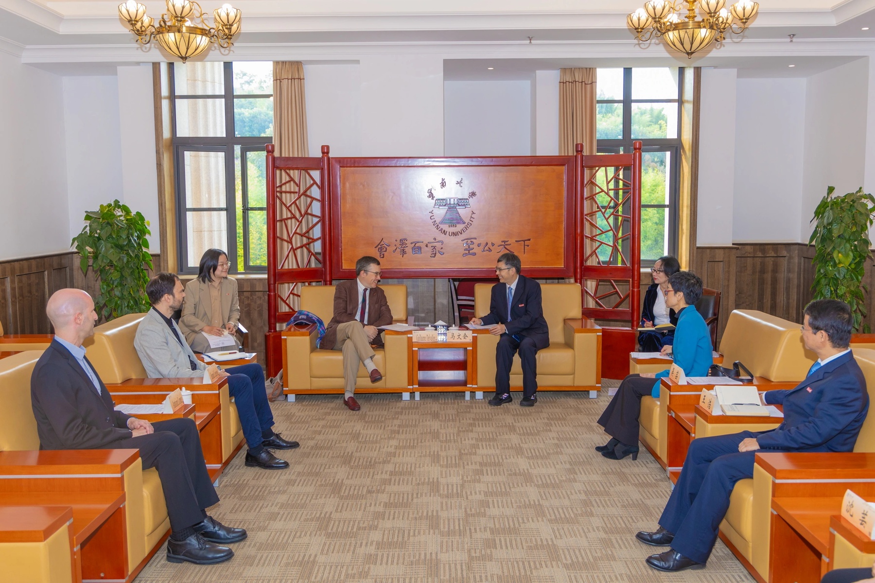 Réunion officielle entre plusieurs personnes assises dans un salon institutionnel lumineux, avec deux interlocuteurs au centre en discussion.