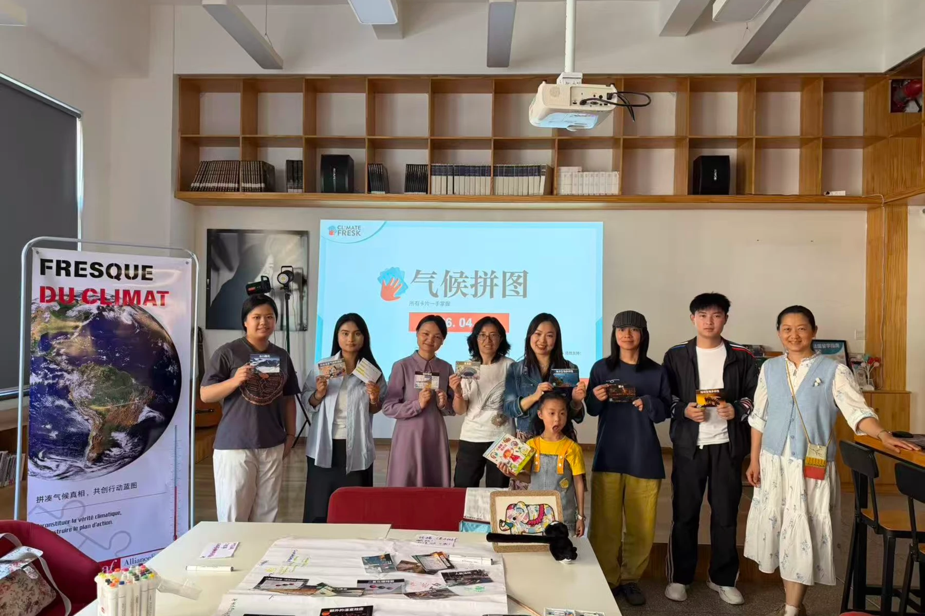 Photo de groupe prise à l’Alliance Française de Kunming à l’issue d’un atelier Fresque du Climat. Plusieurs participants, dont une enfant, posent devant un écran affichant « 气候拼图 » et une bannière « Fresque du Climat », en tenant des cartes de l’atelier.